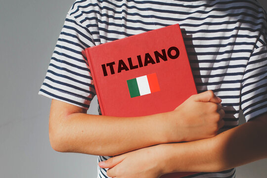 one girl 7 years old schoolgirl in striped t-shirt whose face is not visible holds red italian language textbook with inscription in italian do you speak italian , concept of learning foreign language