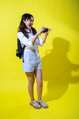 Asian woman with pigtails wearing a white shirt, and a black backpack, holding a camera and smiling enthusiastically, exuding energy and playfulness against a bright yellow background