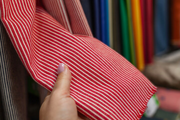 Large selection of natural fabrics in rolls in the store. A woman's hand holds a red striped thick cotton. Close-up.