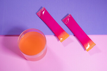 A glass of orange water placed on a pastel purple and pink background, and colored packets,...