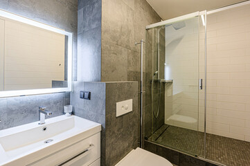interior apartment. Modern bathroom with sleek grey tiles, white vanity, and glass shower enclosure.  Illuminated mirror adds brightness. Minimalist design with chrome fixtures.
