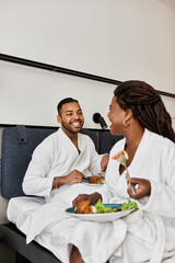 A young couple shares laughter and delicious food while relaxing together in their hotel room.