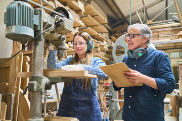 Experienced mentor guiding apprentice in busy lumberyard workshop