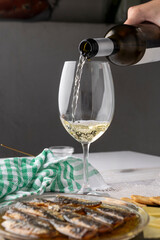A bottle of white wine, a plate of sardines in oil, sliced cheese, and crackers are arranged on a textured mat with a green checkered cloth and a glass nearby.