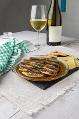 A bottle of white wine, a plate of sardines in oil, sliced cheese, and crackers are arranged on a textured mat with a green checkered cloth and a glass nearby.