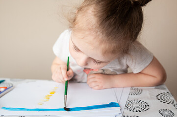 A cute girl draws with paints and pencils at a table near the window.
