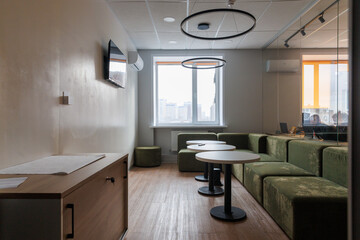 Empty rest area in an open office. White round wooden table surrounded by green sofa. Window on background.