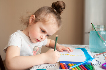 A cute girl draws with paints and pencils at a table near the window.