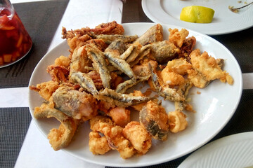 Close-up of white plate filled with a variety mixed of deep-fried seafood, including fish, squid and shrimp. The food is golden brown and crispy with a slice of lemon on a side plate. Street food cafe