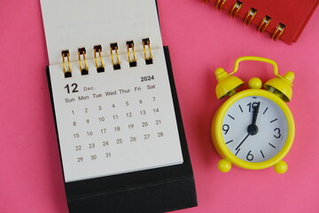Monthly desk calendar for December 2024 year on wooden table with black alarm clock.