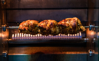 Three whole chickens roasting on a rotisserie in front of a flame grill. The golden-brown, crispy...
