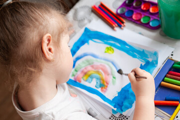 A cute girl draws with paints and pencils at a table near the window.
