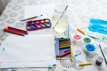 A set of drawing accessories. On the table is a white sheet of paper, watercolor paints, pencils