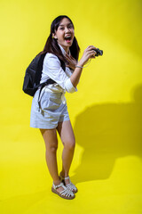 Asian woman with pigtails wearing a white shirt, and a black backpack, holding a camera and smiling enthusiastically, exuding energy and playfulness against a bright yellow background