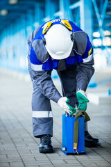 The photo shows a factory worker in a blue suit and a white helmet on his head. He works with his hands at a factory or large enterprise with his head down.