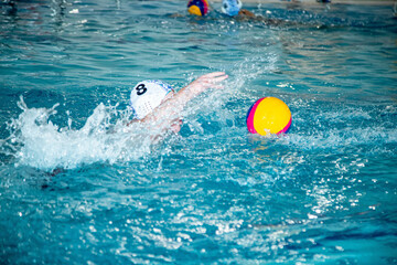 The photo shows children practicing water polo in a large, bright pool.
