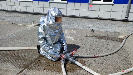 На фотографии спасатель службы мчс в специальном костюме для тушение стольного цвета идет тушить пожар в руках держит ствол с водой. Летний светлый день