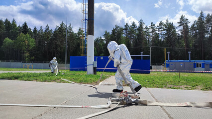 На фотографии спасатель службы мчс в специальном костюме для тушение стольного цвета идет тушить пожар в руках держит ствол с водой. Летний светлый день