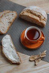 A cozy breakfast setup with slices of rustic bread, one spread with butter, alongside a warm cup of tea in an orange cup on a woven mat