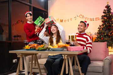 Two handsome young men and two beautiful young women wearing Christmas hats laughing joyfully at a Christmas party with delicious food, drinks and a cheerful exchange of thoughtful gifts