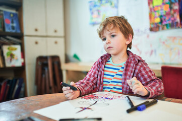 Thoughtful young boy holding markers, sitting at a wooden table with a drawing on paper in an art studio. Shelves and colorful artwork in the background