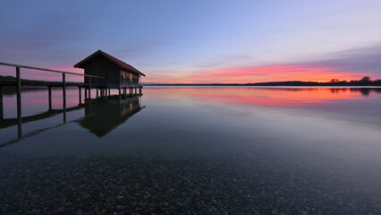 Ein Bootshaus steht einsam im absolut stillen und klaren Wasser eines See. Ein schönes Abendrot zeichnet sich am Himmel ab, das sich im See Spiegelt.