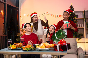 Two handsome young men and two beautiful young women wearing Christmas hats laughing joyfully at a Christmas party with delicious food, drinks and a cheerful exchange of thoughtful gifts