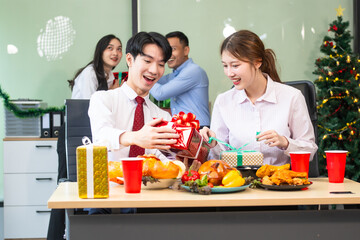 Two young businessmen and two beautiful women sit at an office desk, laughing joyfully while celebrating Christmas.They enjoy food, drinks, and exchange gifts, creating a festive and cheerful office