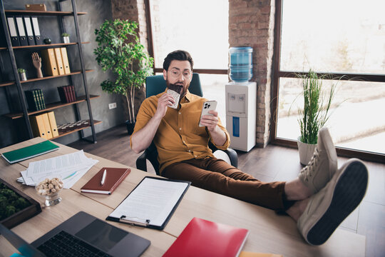 Full length photo of young guy eat chocolate device legs table wear yellow shirt formalwear comfortable startup office loft room interior