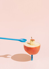 A halved apple rests on a pastel blue screw against a pale pink background. The scene is minimalist, with the colors and simple shapes creating a striking visual contrast. Copy space on the top.