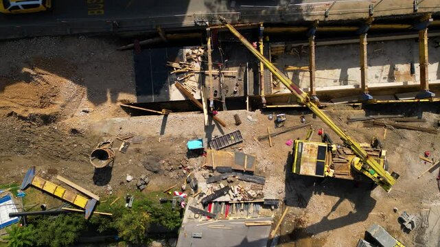 Top-down aerial view of an active urban construction site beside a roadway, with concrete works, formwork, scaffolding, a boom lift and building materials arranged around an unfinished structural sect