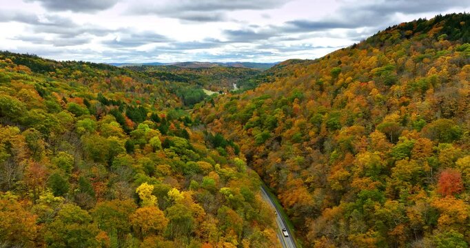 Cars ride by the highway at the foot of the mountains. Aerial view on the rocks wooded with colorful forests in autumn.