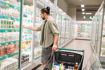 A young man is shopping at the supermarket. A guy walks with a cart between refrigerators with dairy products in a large store