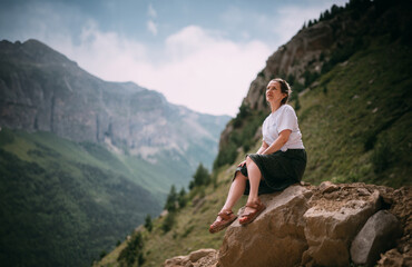 Naklejka premium Portrait of a female tourist sitting on a rock against the background of mountains. A young girl in a dress meditates, sits in peace high in the mountains