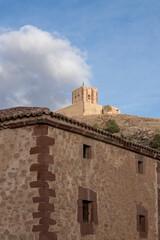 Naklejka premium Historic stone building with rustic windows and a medieval castle tower on a hill under a cloudy blue sky.