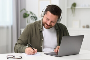Fototapeta premium Interpreter in headphones taking notes while having video chat via laptop at white table indoors