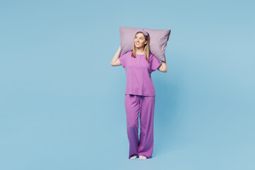 Full body calm young woman wear purple pyjamas jam sleep eye mask rest relax at home hold in hand pillow behind head look aside on area isolated on plain blue background. Good mood night nap concept.