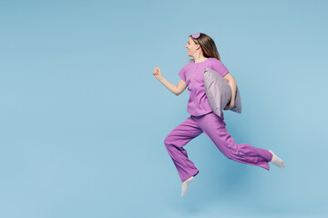 Obraz premium Full body calm happy young woman wear purple pyjamas jam sleep eye mask rest relax at home jump high hold pillow run fast hurry up isolated on plain blue cyan background. Good mood night nap concept.