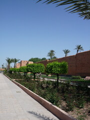 Ancient clay ramparts of Marrakech stretch along a landscaped garden path, lined with palm trees and metal railings.
