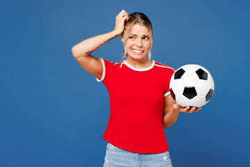 Young sad woman fan wearing basic red t-shirt cheer up support football sport team hold in hand soccer ball watch tv live stream shrugging shoulders looking puzzled isolated on plain blue background.