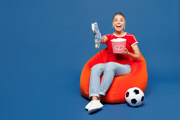 Full body young woman fan wear red t-shirt foam 1 fan glove finger up support football sport team hold soccer ball watch tv live stream sit in bag chair eat popcorn isolated on plain blue background.