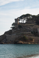 Close-up of a historic coastal fortress perched on a rocky hill, overlooking the turquoise sea, with winding stone walls and silhouetted trees against a soft blue sky.
