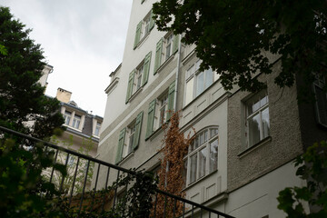 A beautifully designed urban building with decorative green shutters and lush ivy climbing up its walls, set against a serene backdrop of a cloudy sky within a charming city environment