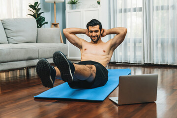 Athletic and sporty man doing crunch on fitness mat while follow online home workout exercise instruction for fit physique and healthy sport lifestyle at home. Online gaiety home exercise video.