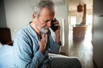 Senior man talking on the smartphone holding chest at home