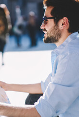 Fototapeta premium Cheerful bearded tourist in sunglasse resting outdoors on street of architectural downtown.Positive young man traveler in spectacles holding map in hands to find showplaces during trip