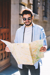 Positive bearded tourist happy to find right location of architectural showplaces on map strolling in uran setting in sunny day.Cheerful male traveler in stylish sunglasses walking on streets © BullRun