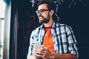 Positive bearded blogger in eyeglasses for vision correction laughing during friendly messaging in online chat on smartphone via 4G internet connection.Cheerful young man sending sms on cellular