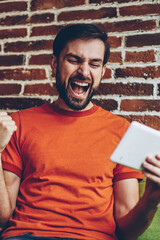 Overjoyed bearded young man celebrating victory of favourite team watching live stream online on website on modern tablet device connected to 4G internet.Excited hipster blogger got win on web page