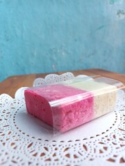 Two layers of red and white sweet cake, wrapped in plastic, on a white base, on a wooden table, blue background.